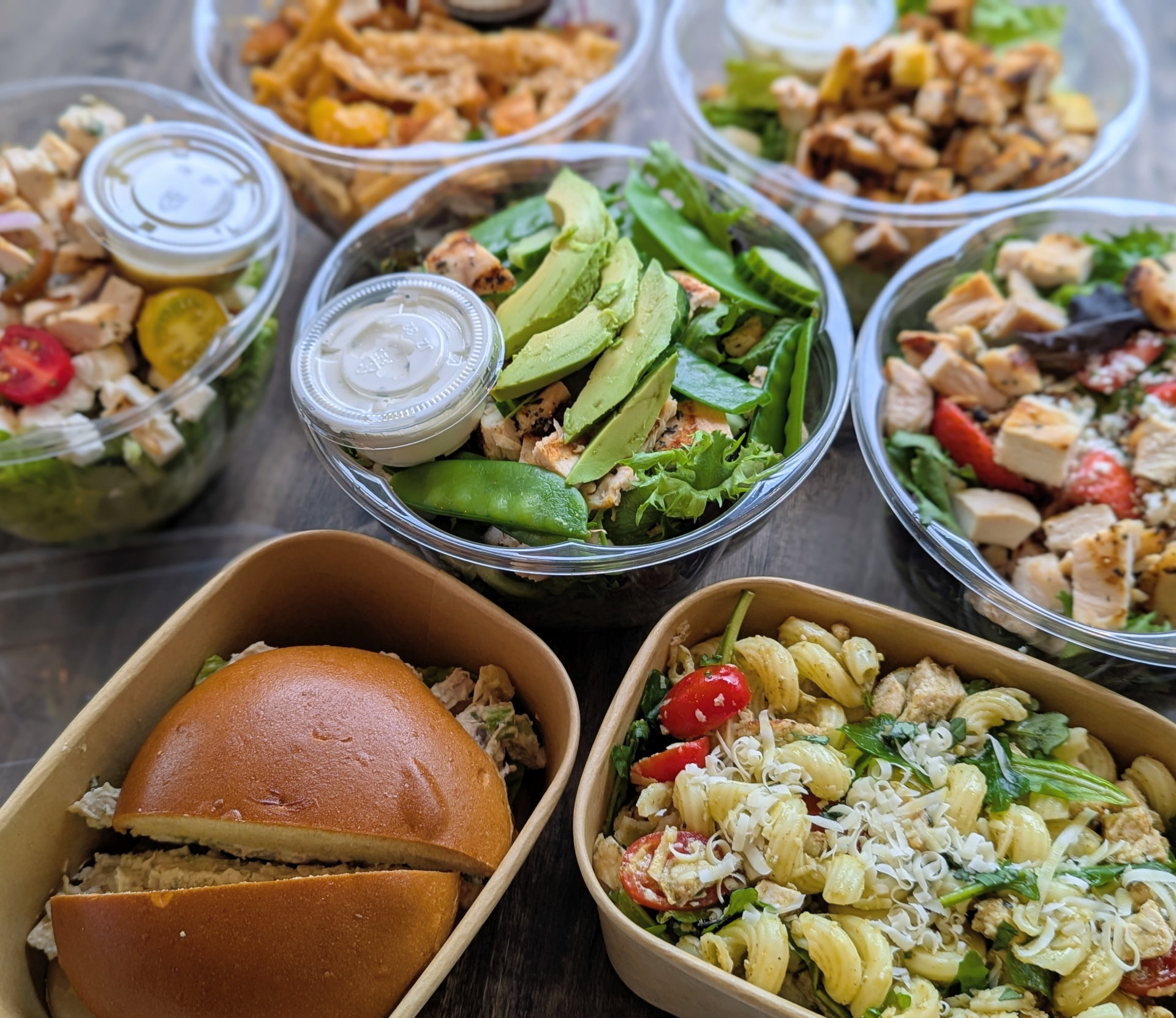 Catering packed to go: a sandwich, pasta salad and grilled chicken salads in containers on a wood table